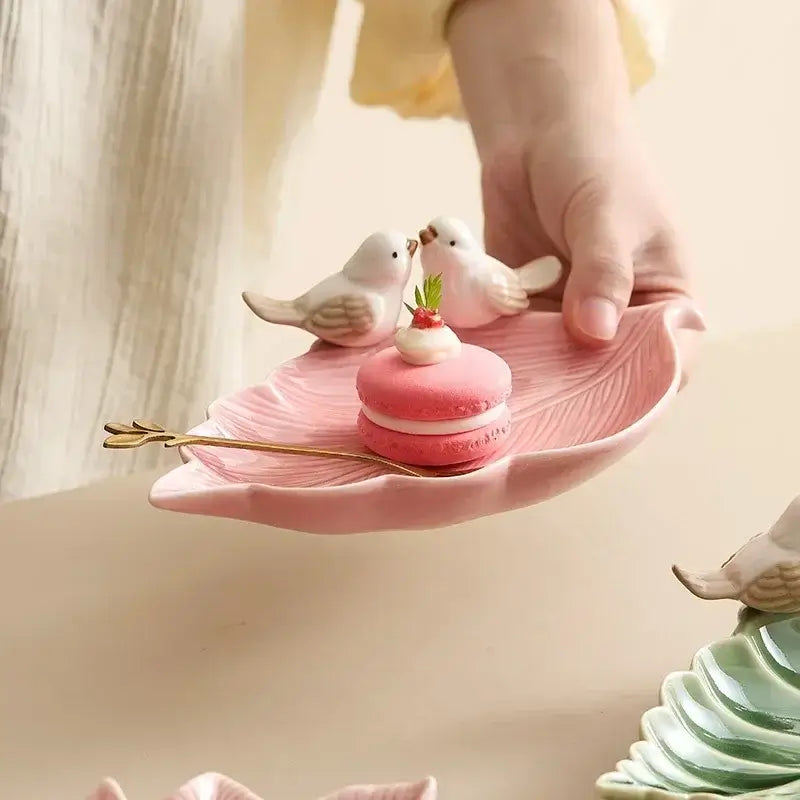 Hand holding a pink ceramic leaf jewelry tray with bird figurines, showcasing its use for storing small accessories or treats.