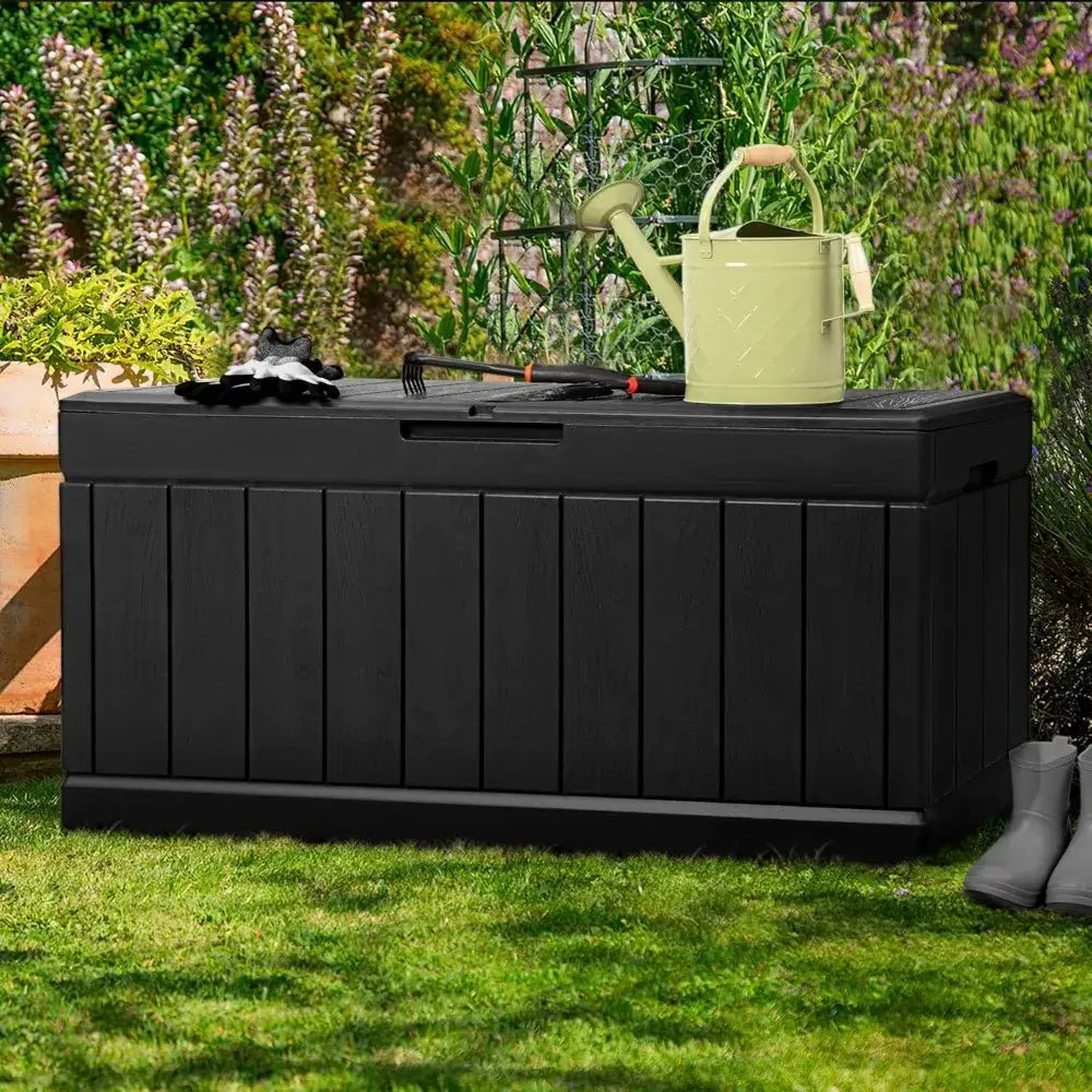 Black 83 Gallon Outdoor Deck Box used in garden setting with tools and watering can on top. Durable resin construction for outdoor use.